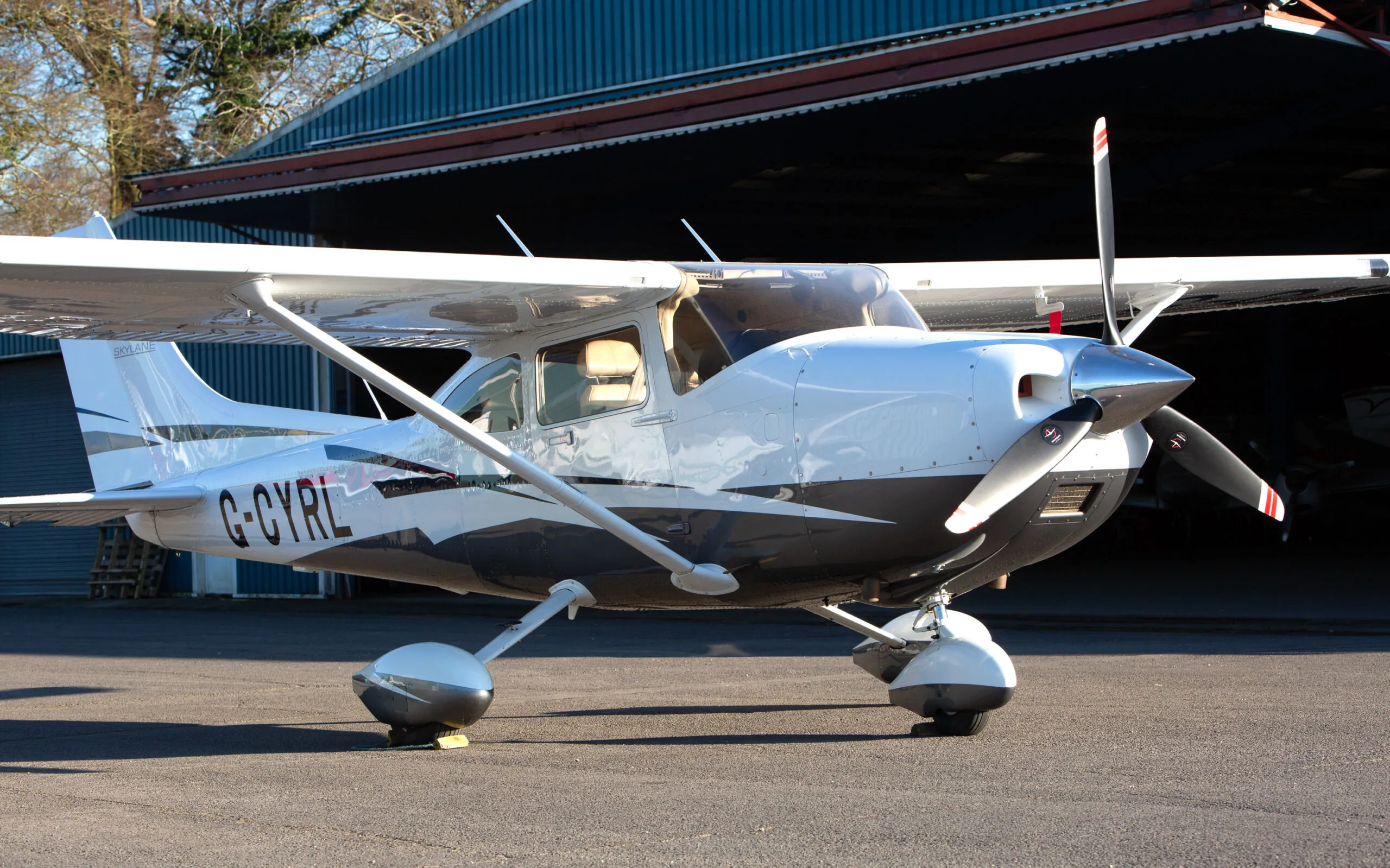 A white and black Cessna 182T (G-CYRL) parked on an airfield with a hangar in the background.
