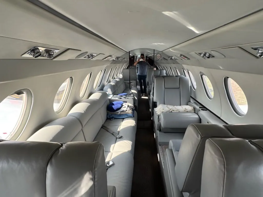 Long view of a business jet cabin interior with light gray leather seating, including a sofa, and a person standing in the rear.