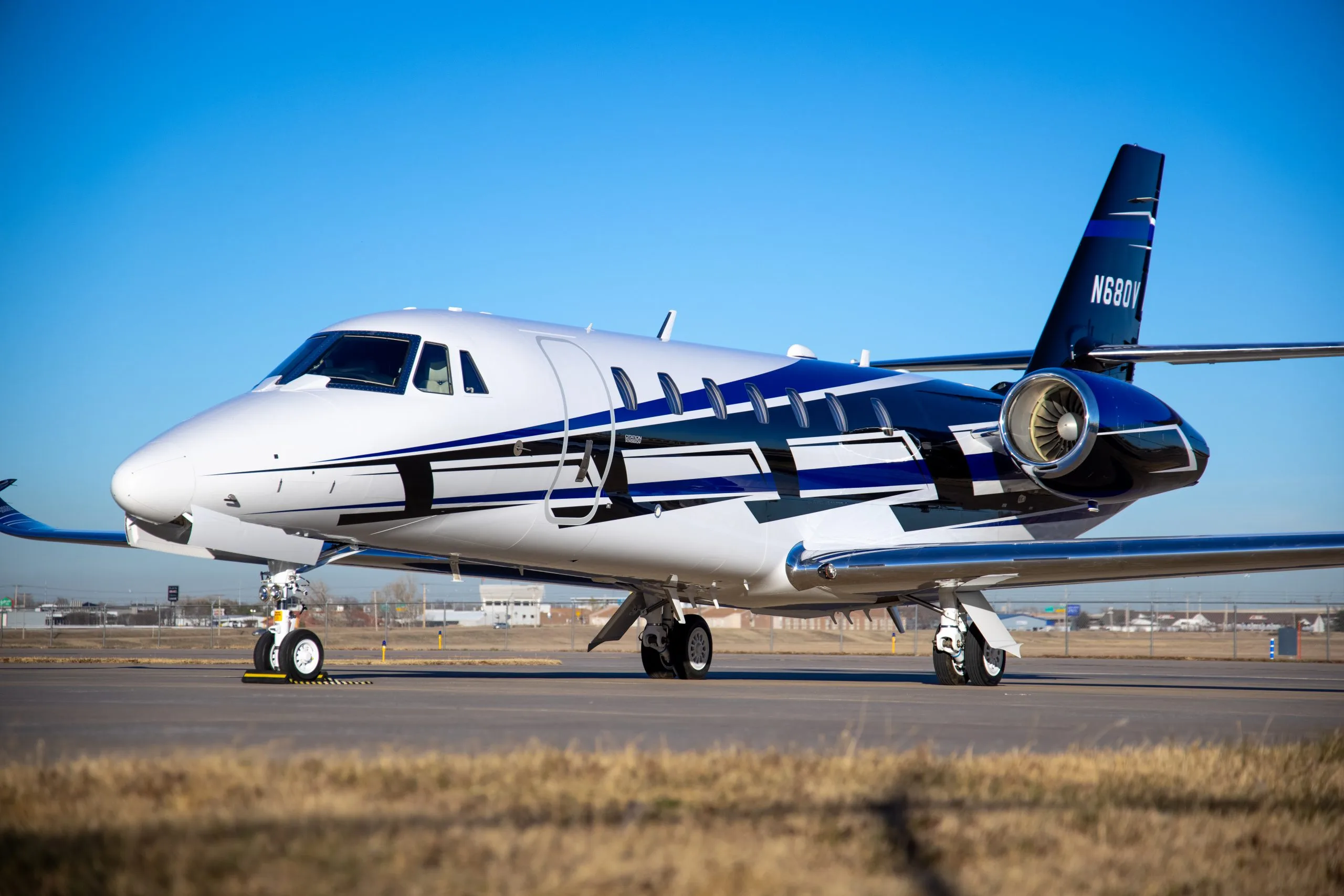 A white Cessna Citation XLS+ business jet with a bold blue and black stripe design, parked on an airfield on a sunny day.