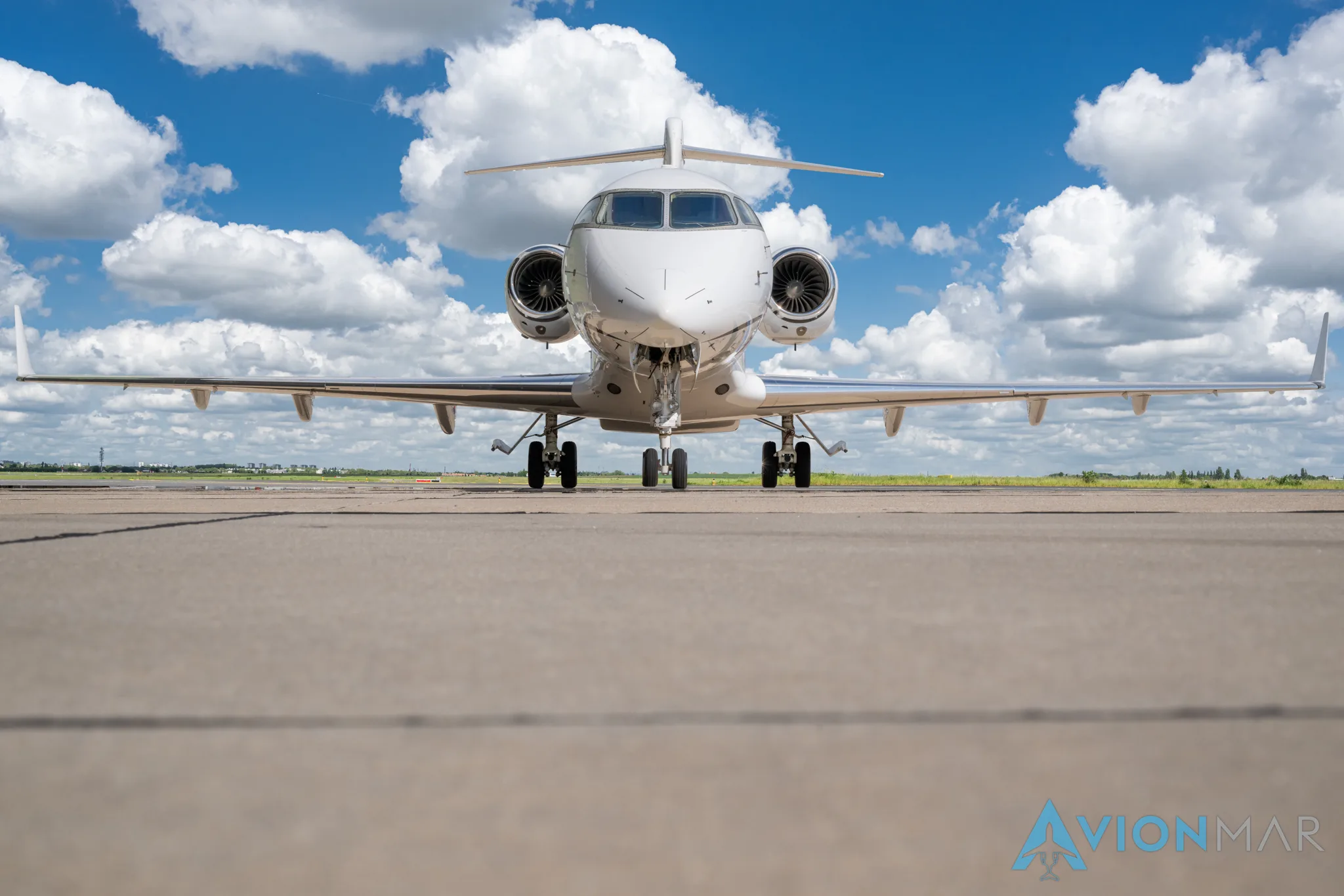A white Bombardier Challenger 350 business jet on the tarmac under a blue sky with clouds, viewed from the front.