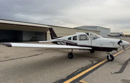 A white and maroon single-engine propeller aircraft, N4796S, parked on an airfield tarmac with hangars in the background under an overcast sky.