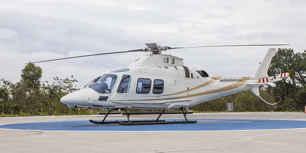 A white helicopter with gold stripes is parked on a blue helipad under a cloudy sky, viewed from the side.