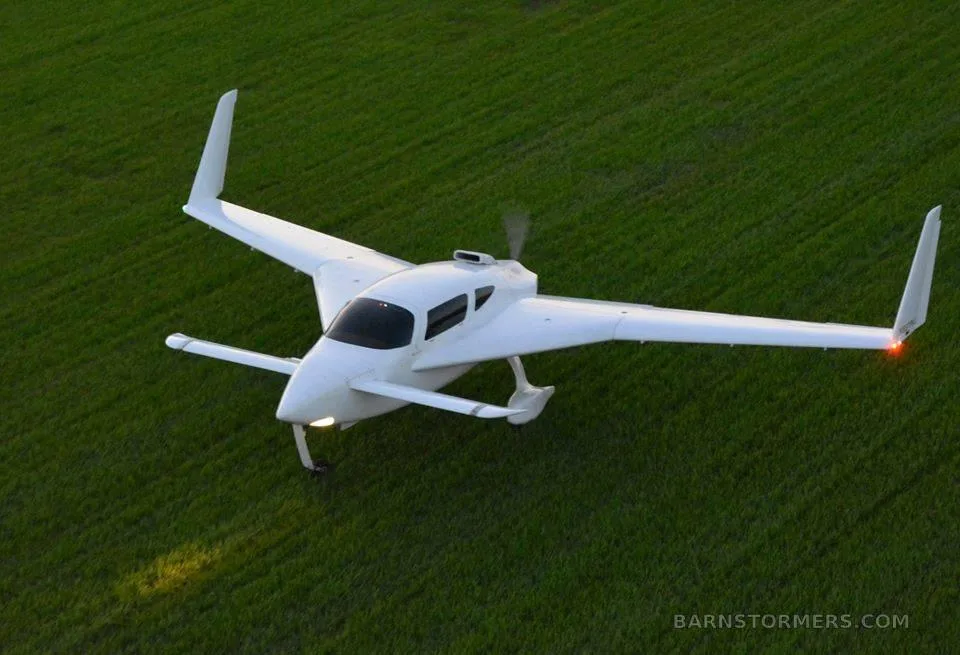 A white, single-engine, pusher-propeller aircraft with twin tail booms flies low over a green field.