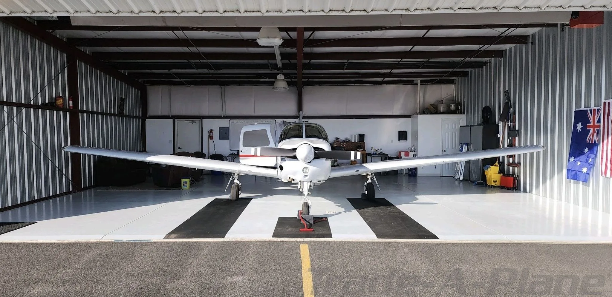 A white Piper aircraft with a black propeller is parked head-on inside a metal hangar.
