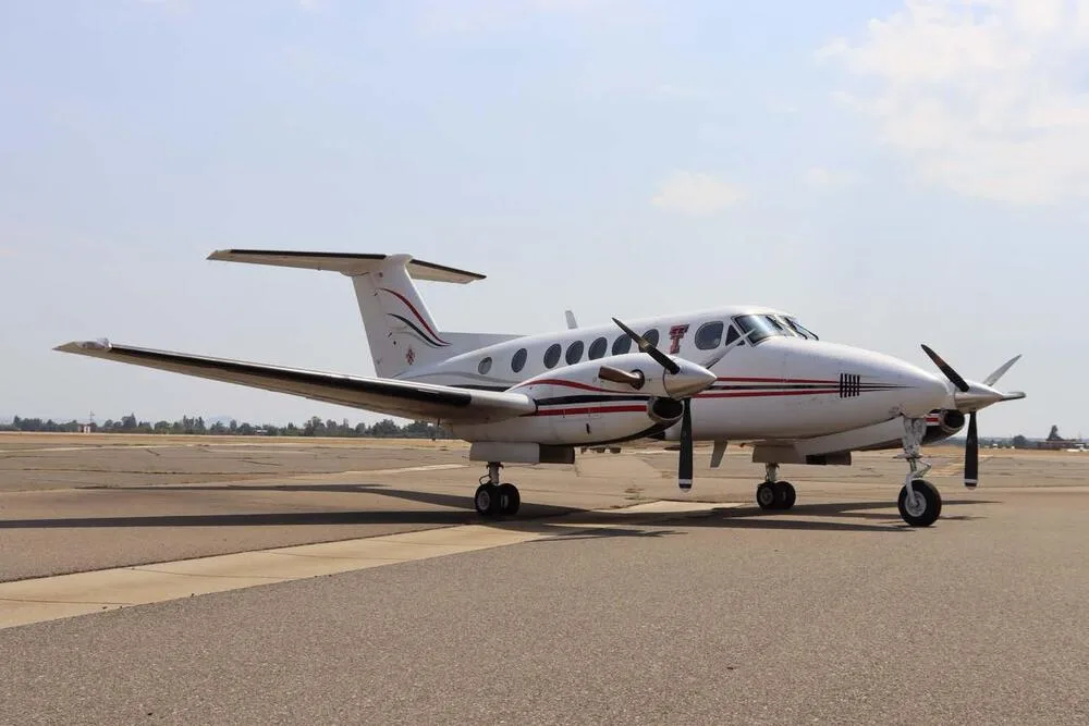 A white Beechcraft King Air with red and black stripes, viewed from the front-side on an airport tarmac.