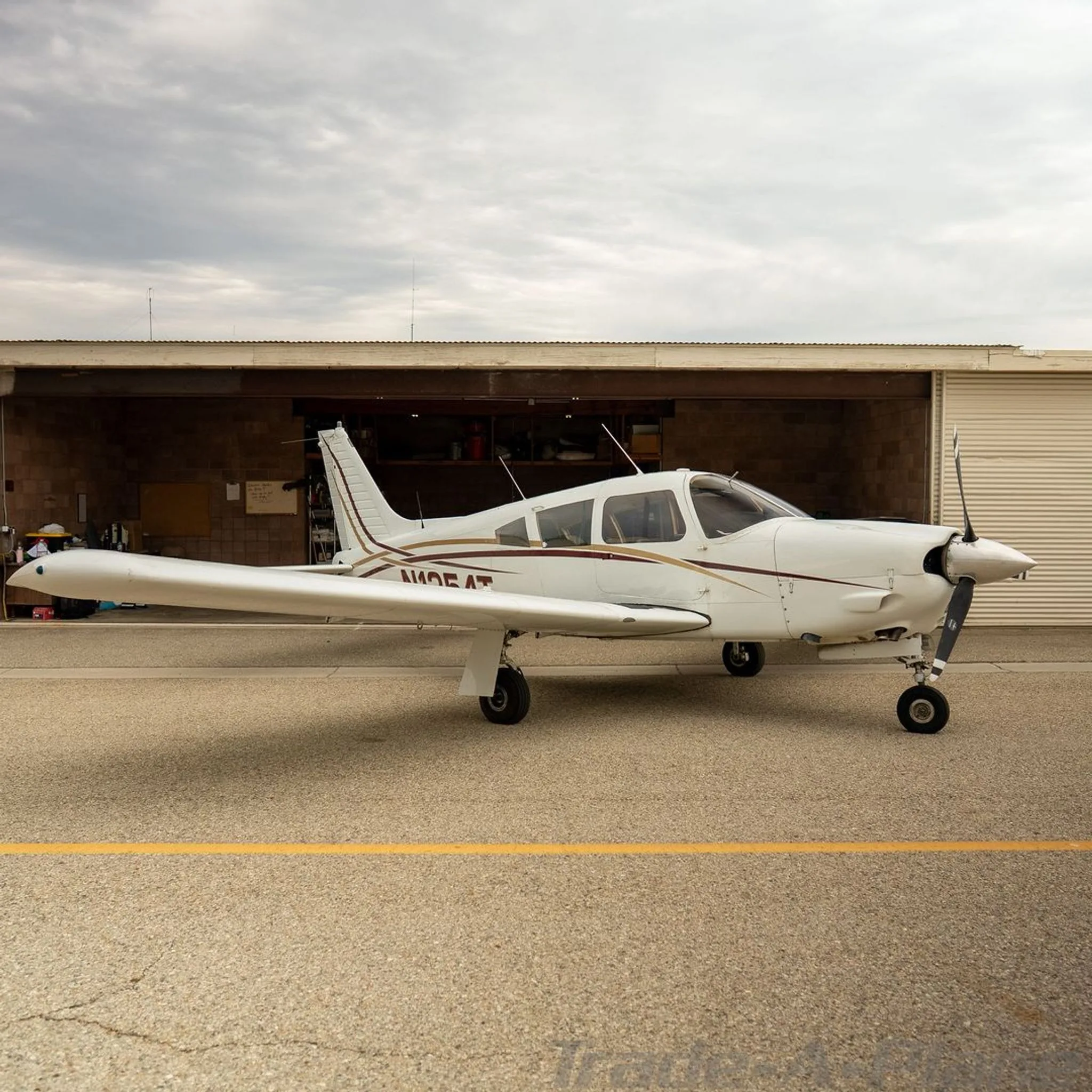 White Piper Cherokee N1354T with maroon and gold stripes parked on an airfield next to a hangar.