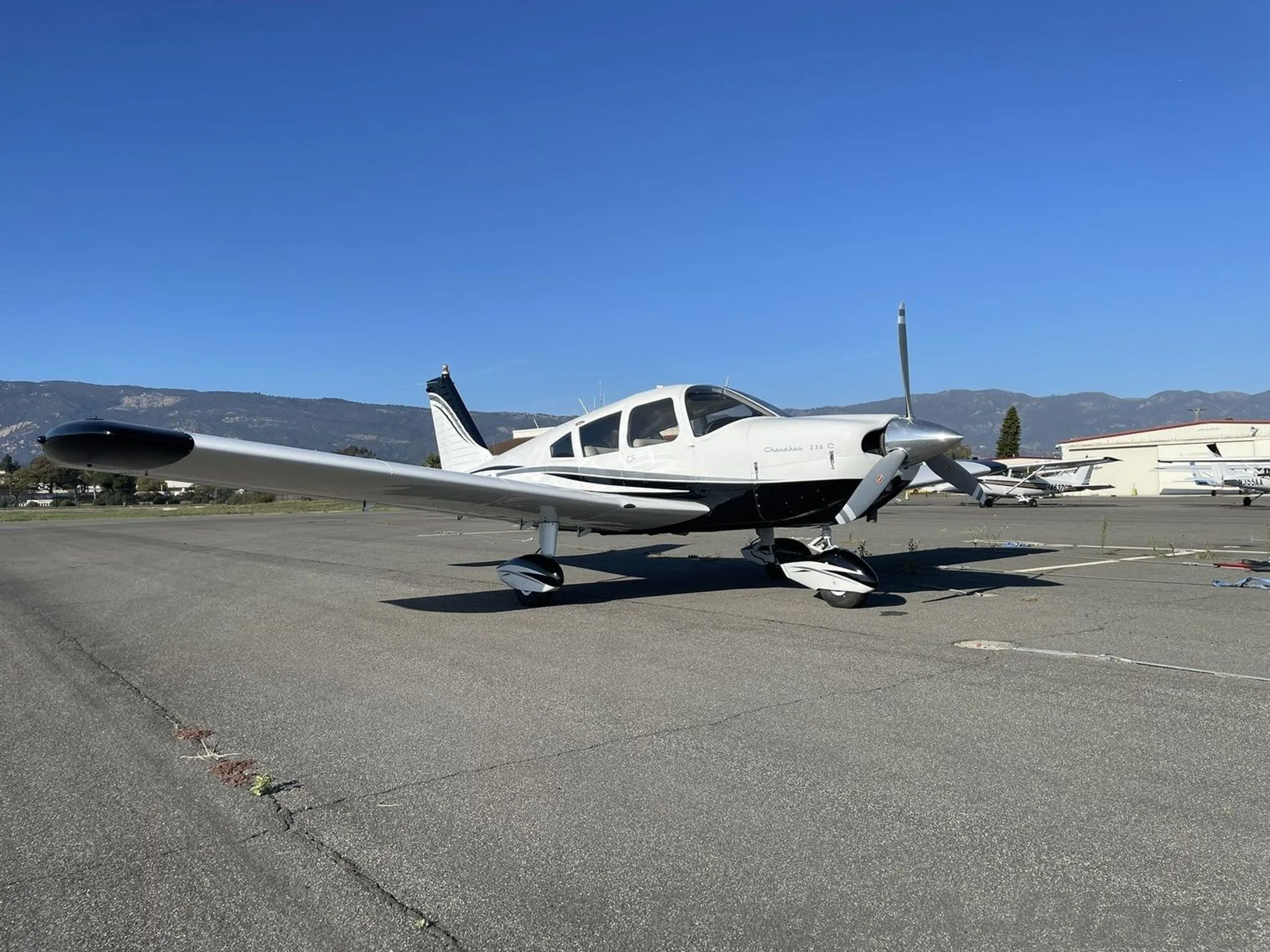 A front-side view of the white and black Piper Cherokee 235 C aircraft N991CB parked on an airfield with mountains in the distance.