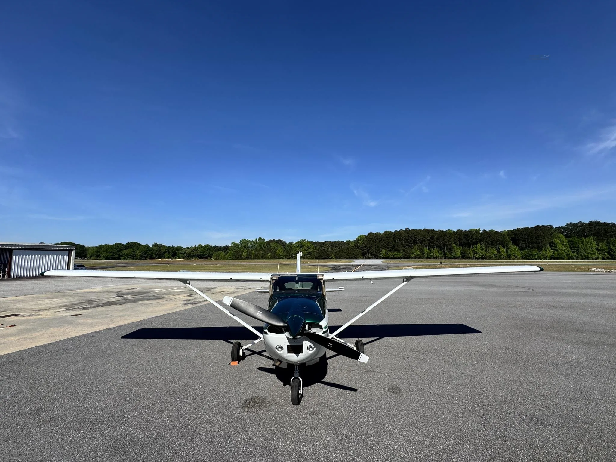 A head-on view of the white and green Cessna 182 Skylane, N49FC, parked on an airfield with trees in the background.