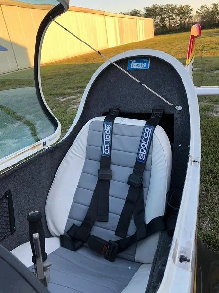The interior of a small aircraft showing a grey and white seat with a Sparco harness and an open canopy.