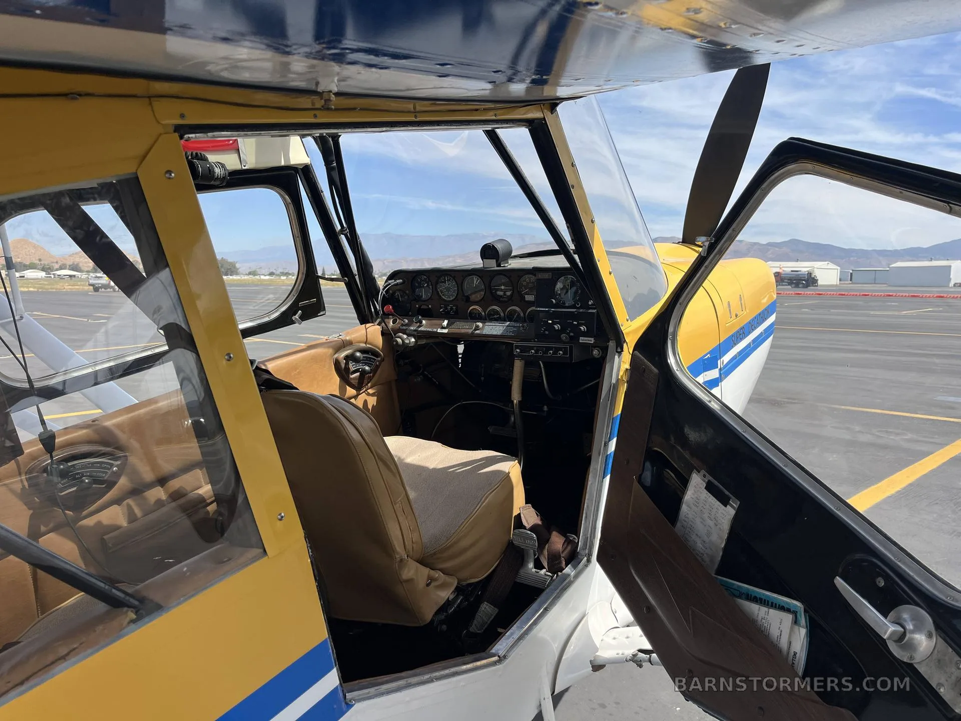 View into the cockpit of a yellow aircraft, showing the instrument panel, brown seats, and open door with an airport in the background.