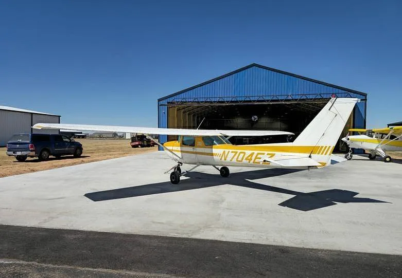 A white and yellow Cessna 150, registration N704EZ, parked on a tarmac in front of a blue hangar.