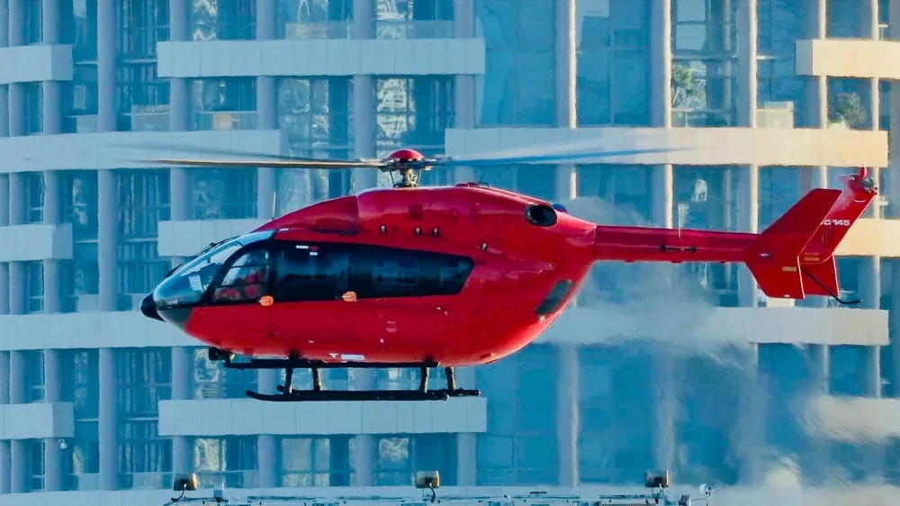 A red Airbus H145 helicopter hovers in front of modern glass buildings.