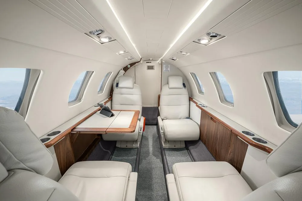 The luxurious interior cabin of a private jet with light leather seats, a wooden table, and large windows.