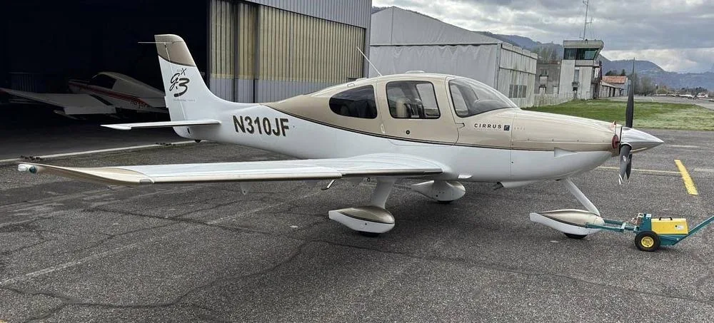 A Cirrus SR22 G3 aircraft with a two-tone tan and white paint scheme is parked on an airfield tarmac with a tug attached to the nose wheel.