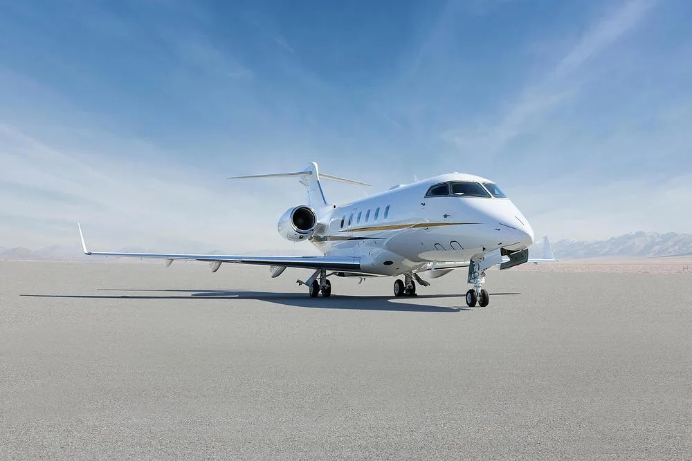A white Bombardier Challenger 350 business jet with gold and black accent stripes parked on a tarmac, viewed from the front-right.