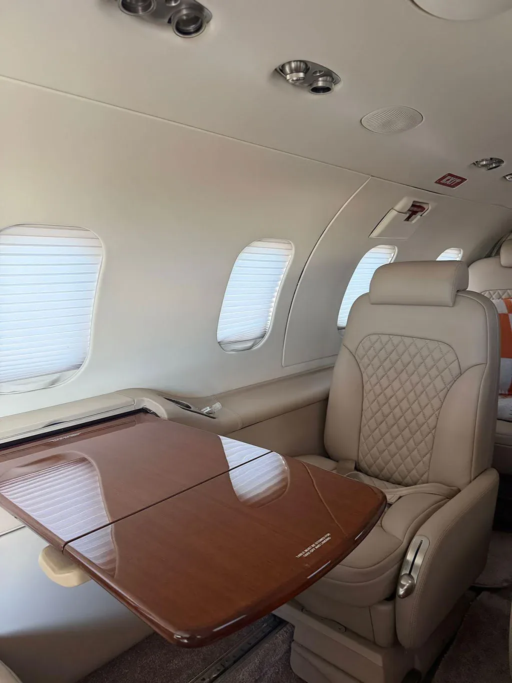 The aircraft cabin features a fold-out wooden table next to a light beige quilted leather seat.