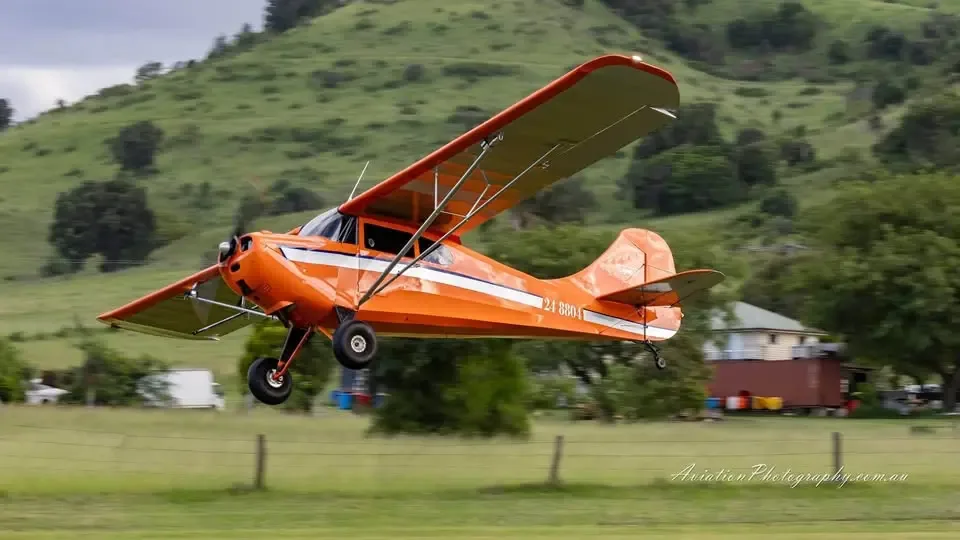 An orange and white high-wing aircraft with tail number 24-8804 flying low over a green field with hills in the background.