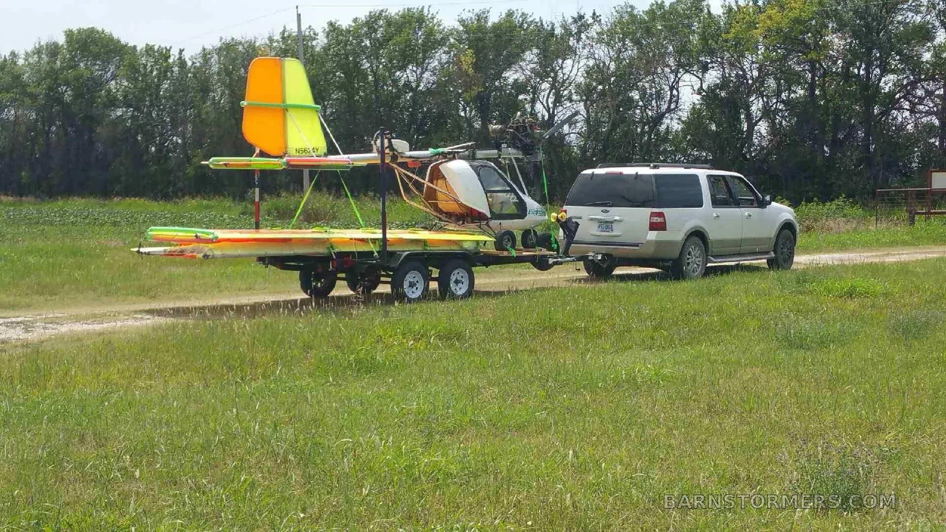 A disassembled ultralight aircraft with orange and yellow wings loaded on a trailer, being towed by a white SUV on a grassy dirt road.