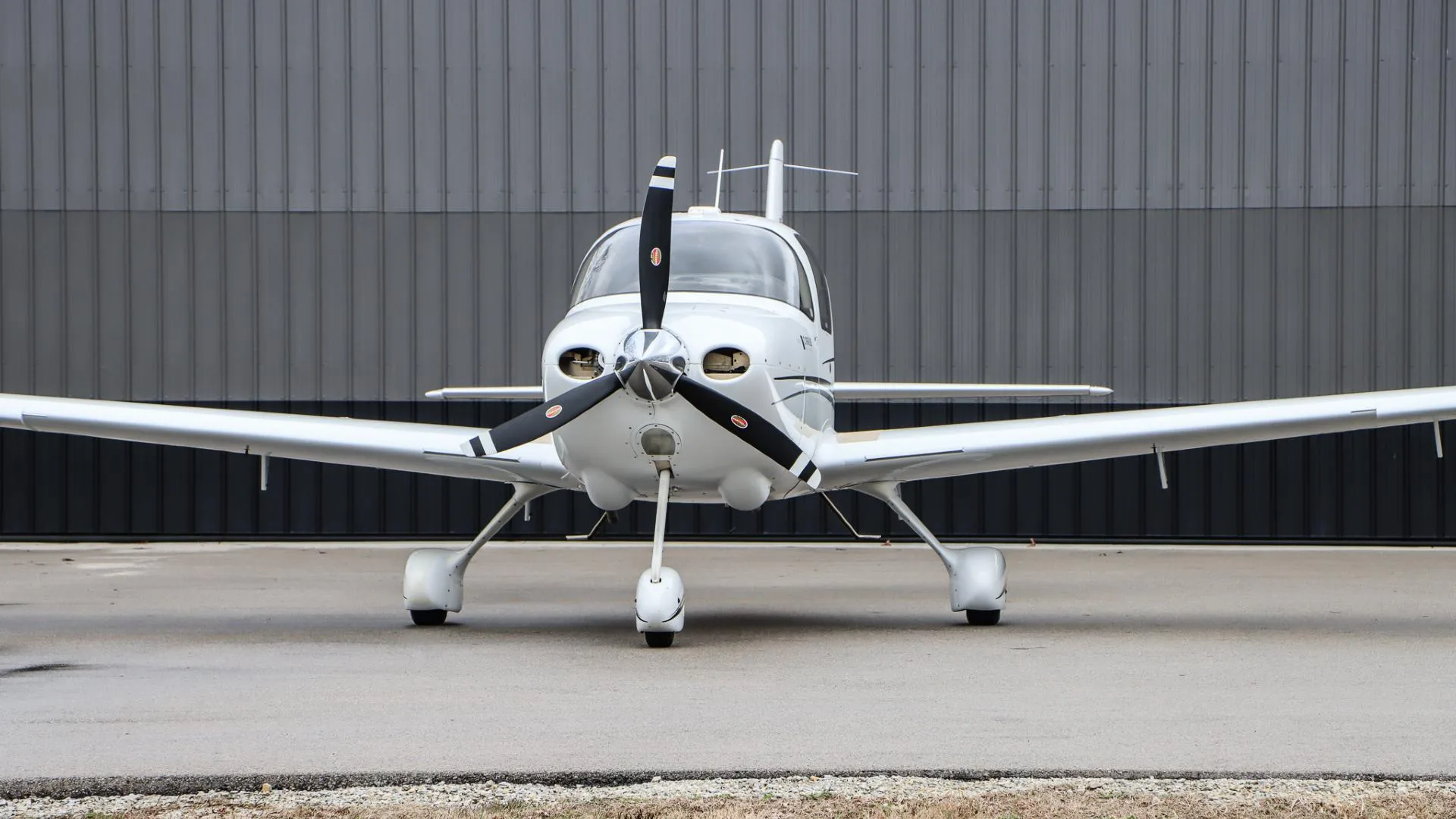 A white Cirrus SR20 GTS aircraft, N984PG, is parked head-on against a dark gray hangar wall.