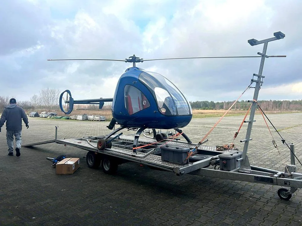 A blue single-seat helicopter is secured on a transport trailer on an airfield, with a person walking nearby under a cloudy sky.