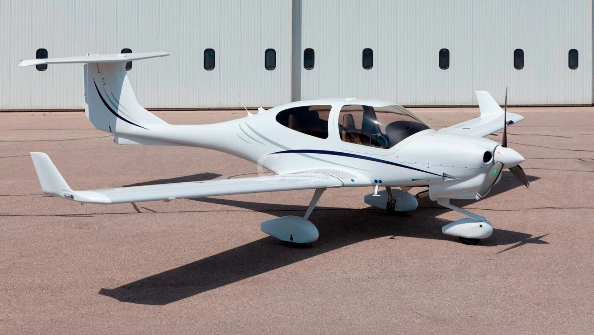 A white Diamond DA40 aircraft with blue and grey stripes parked on an asphalt apron in front of a hangar.