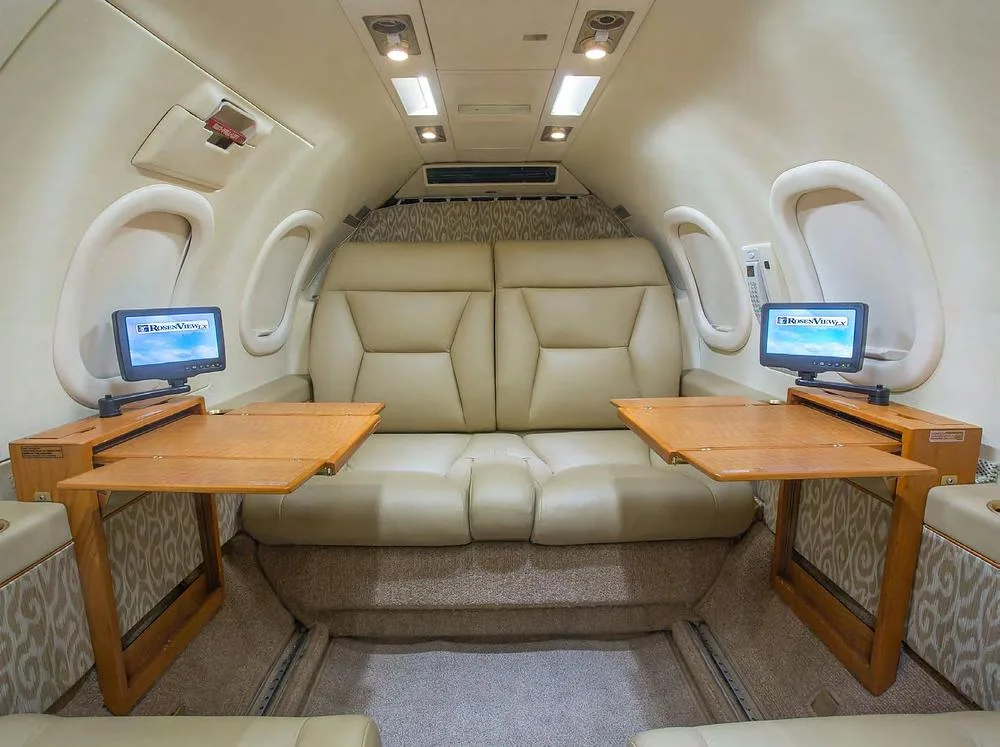 A view of the aircraft cabin interior with two beige leather seats, fold-out wooden tables, and small entertainment monitors.