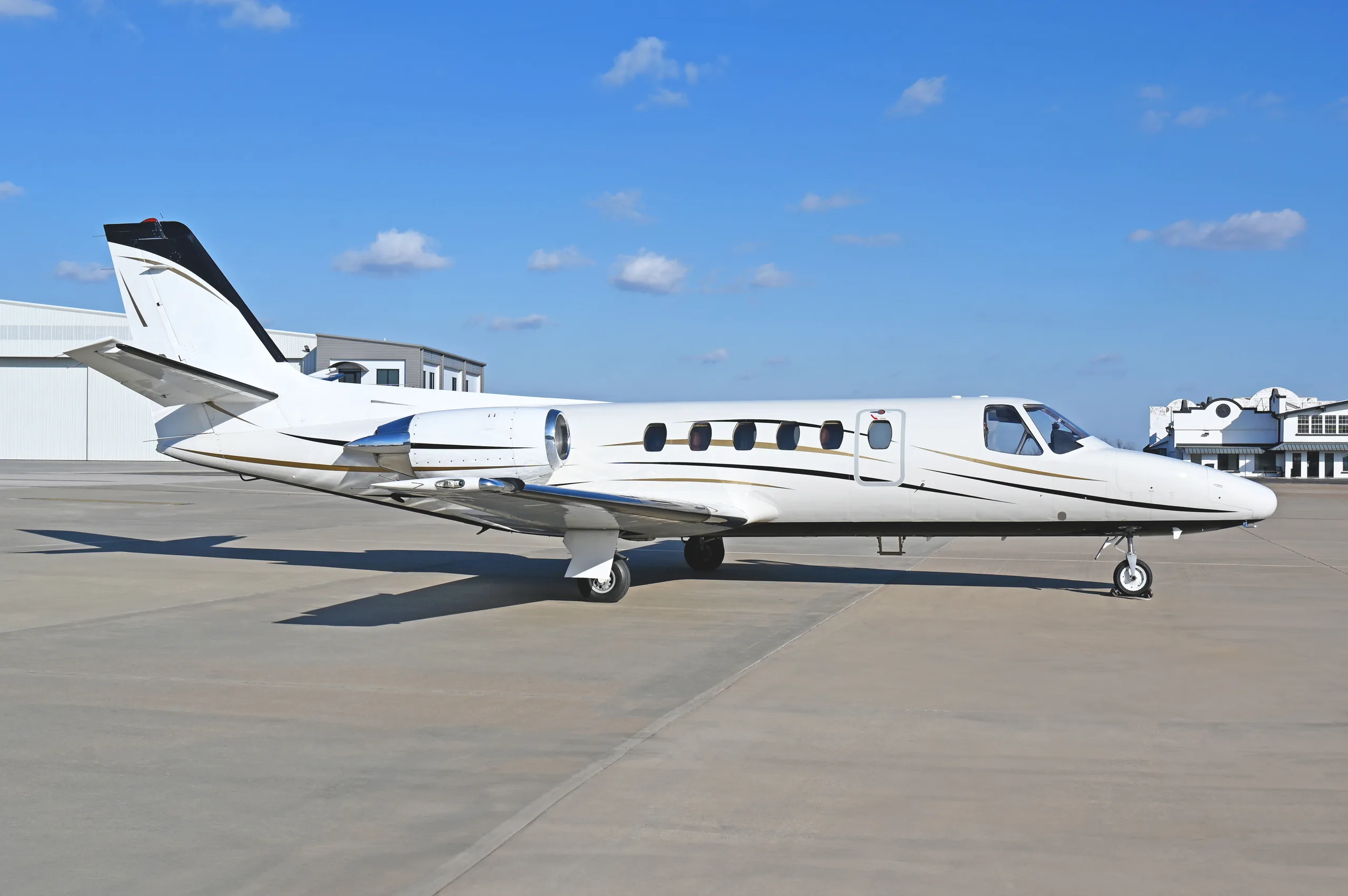 A white business jet with black and gold stripes parked on an airfield tarmac under a blue sky.