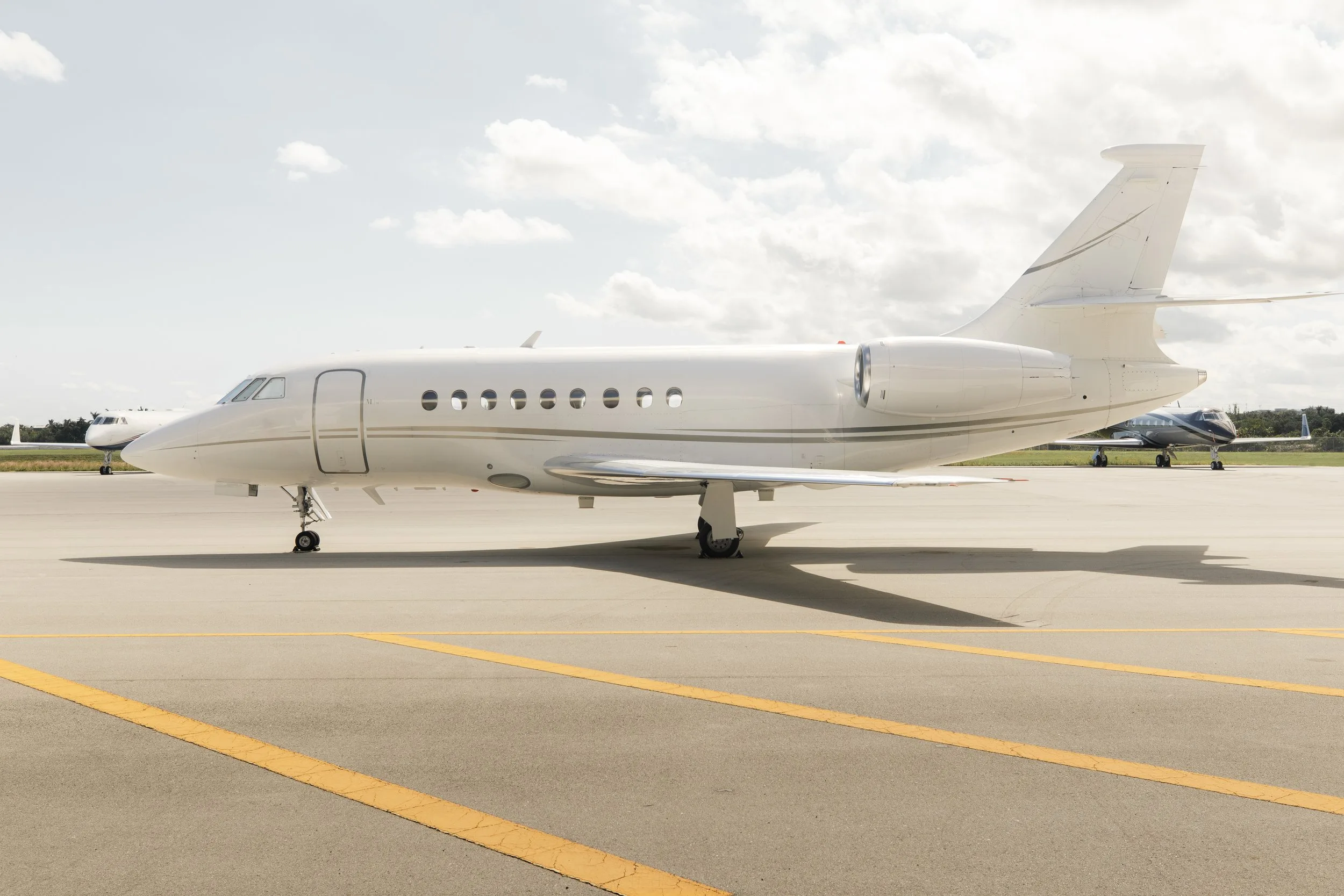 A white Dassault Falcon business jet with gold accents on the tarmac under a partly cloudy sky.