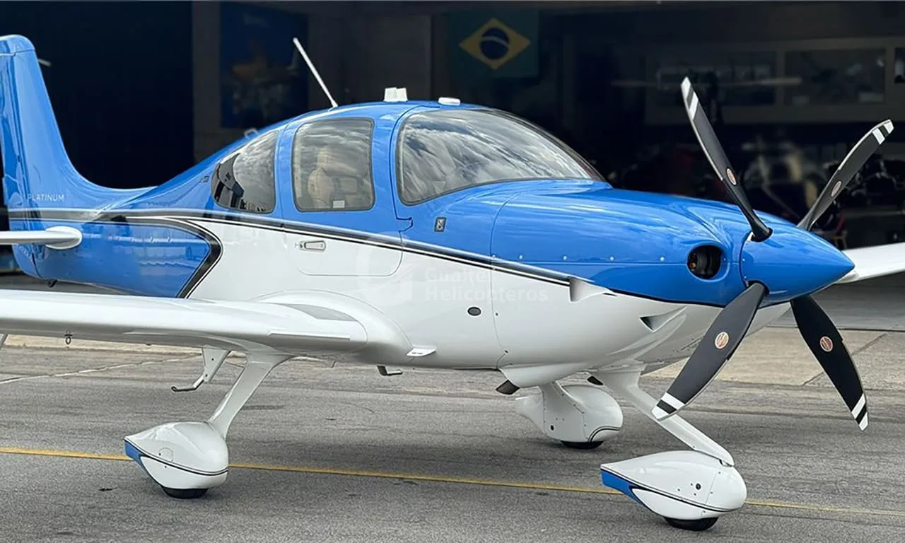 A blue and white Cirrus SR22 aircraft parked on an tarmac with a hangar and Brazilian flag in the background.