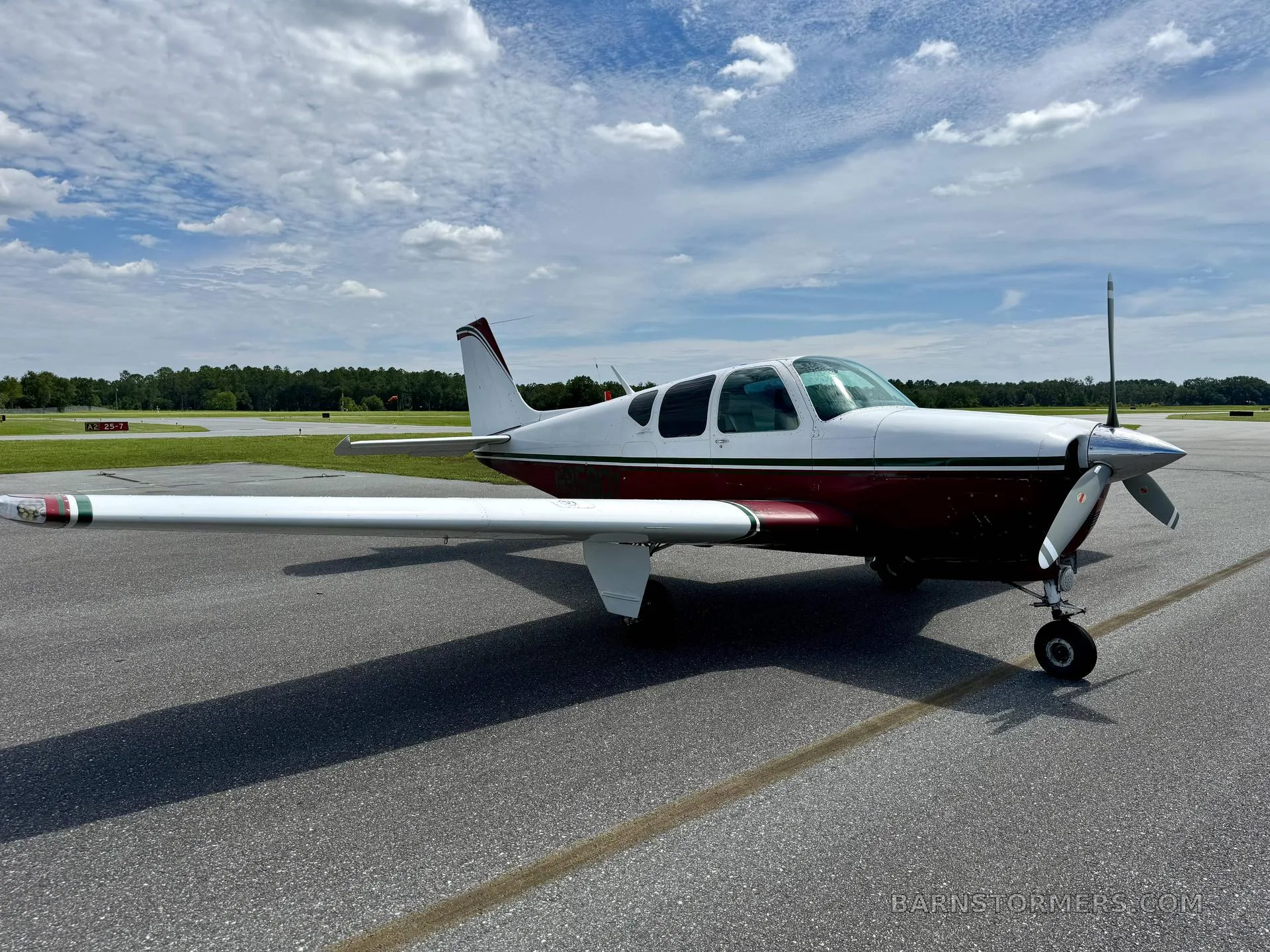 A white and maroon single-engine propeller aircraft is viewed from the front-right on an airfield under a cloudy sky.