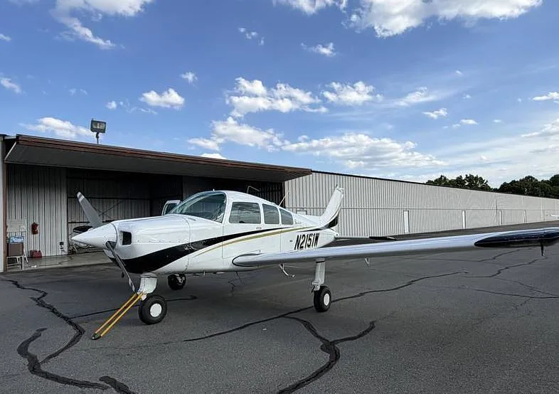 A white and black single-engine propeller aircraft, N2151W, is parked on an asphalt ramp in front of a hangar under a partly cloudy sky.