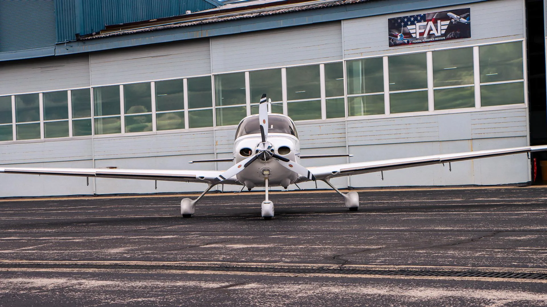 A white Cirrus aircraft with a three-bladed propeller is parked on a tarmac in front of a hangar.