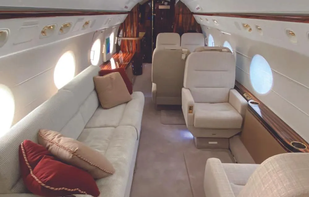 A view of the private jet's cabin, showing a cream-colored sofa with red and brown pillows, and individual cream seats.