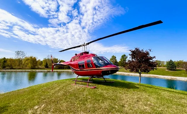 A red Bell 206 JetRanger helicopter with registration N41PH sits on a grassy mound next to a pond under a blue sky.