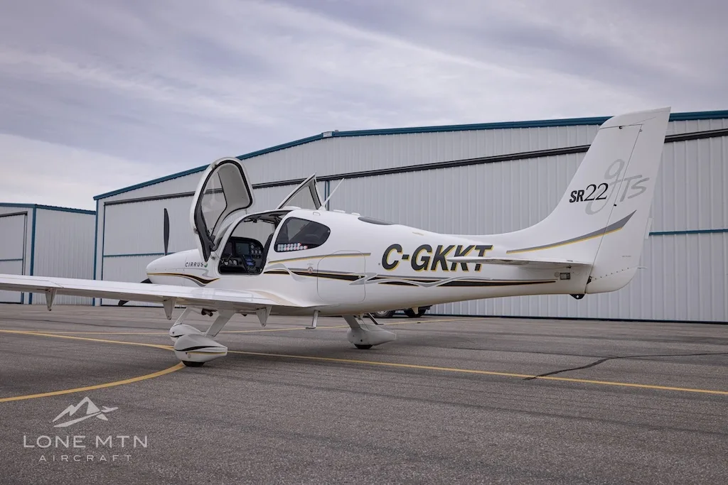 A slightly different rear three-quarter view of a white Cirrus SR22 aircraft with registration C-GKKT, doors open, on an airfield.