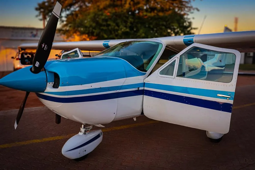 A white and blue Cessna 210 Centurion aircraft with its door open, parked on a tarmac at sunset.