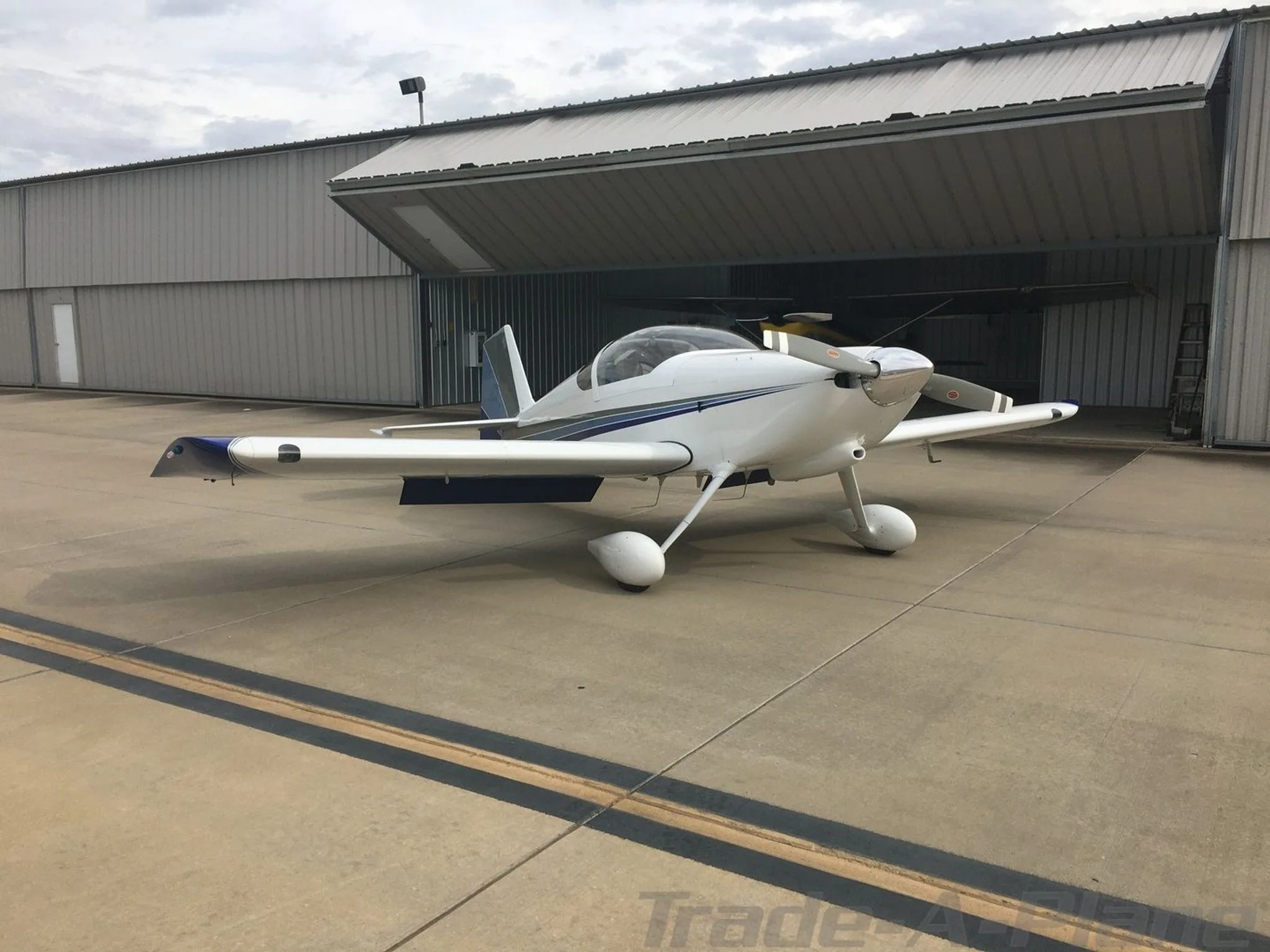 A white Van's RV-6 aircraft with blue and gray stripes parked on an airfield in front of an open hangar.