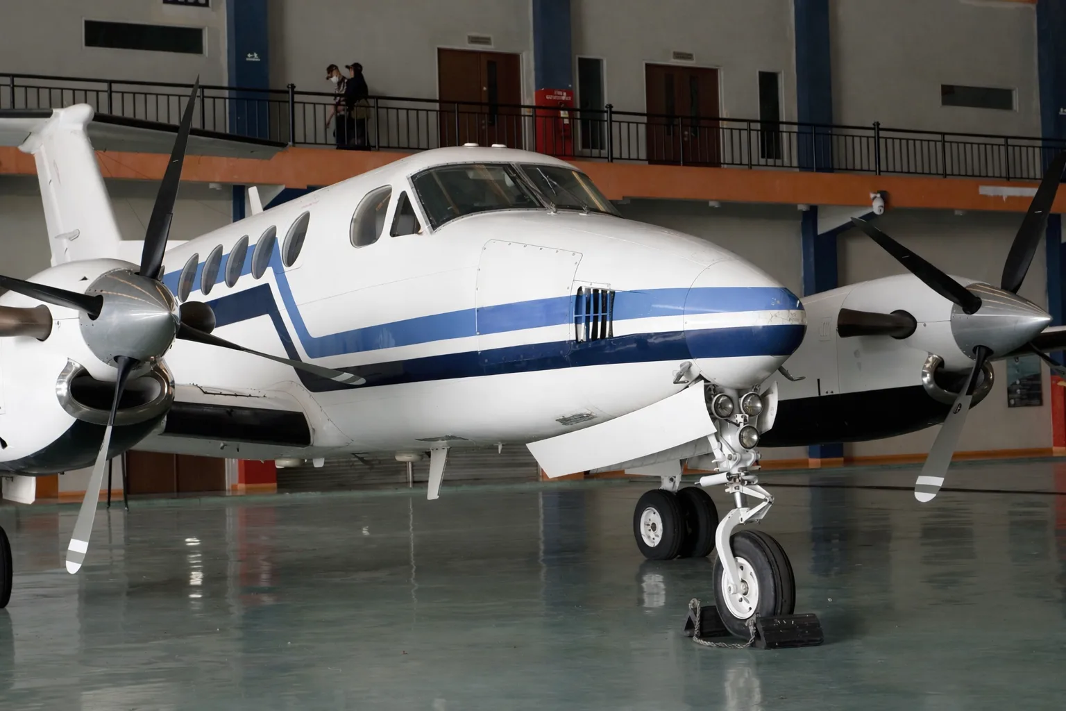 A close-up view of the front of a white and blue twin-turboprop aircraft, showing its nose and propellers, inside a hangar.