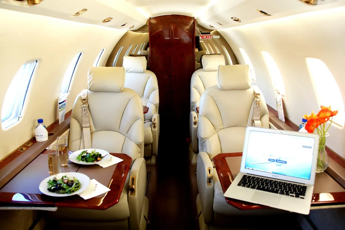 The luxurious interior of a private jet cabin shows multiple beige leather seats, tables with food, and a laptop.