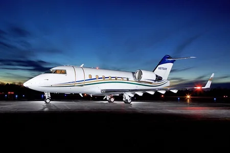 A white business jet with blue, green, and gold stripes and a blue tail, parked on an airfield at dusk.