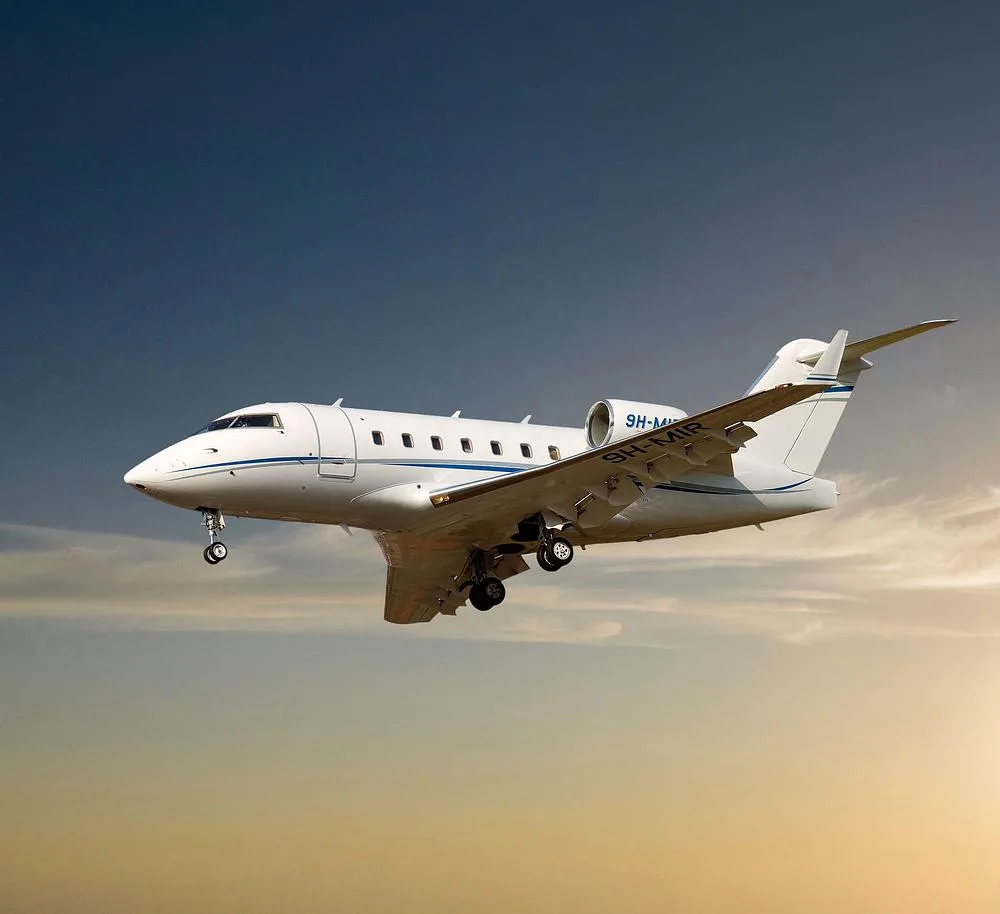 A white Bombardier Challenger 605 business jet with blue stripes, registration 9H-MIR, flying at sunset with landing gear extended.