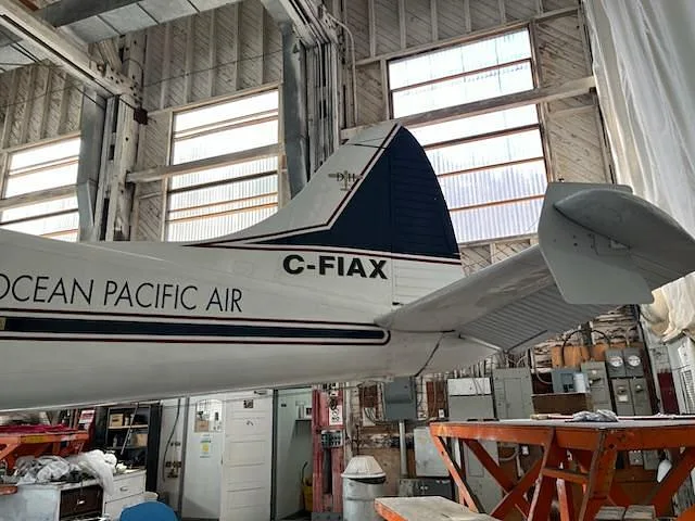 The tail section of a white De Havilland Beaver seaplane with "C-FIAX" markings is shown inside a hangar with large windows.