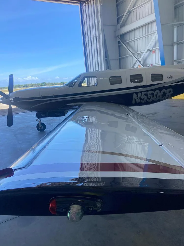 A close-up view of the wing and fuselage of the Piper M500 aircraft N550CR, with the hangar door open to a sunny sky.