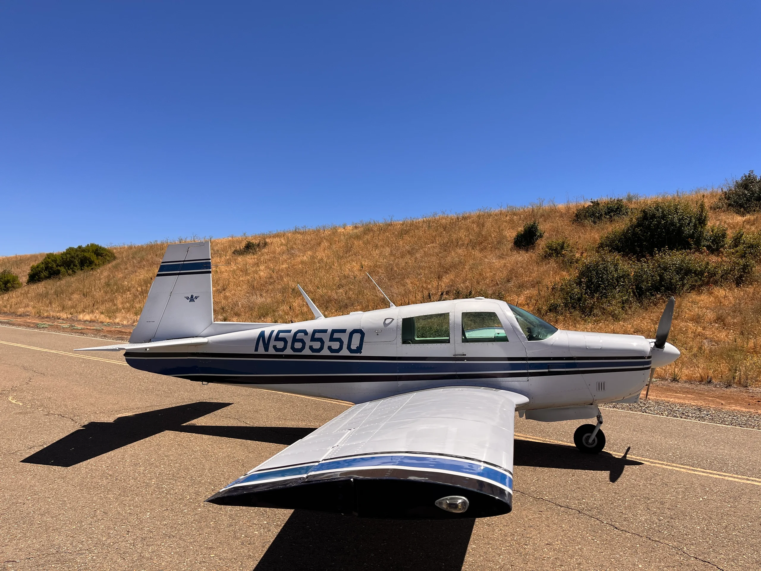 A white single-engine propeller aircraft with blue stripes and tail number N5655Q is parked on an asphalt surface with a dry hill in the background under a clear blue sky.