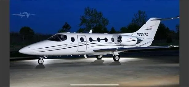 A white business jet, N224FD, with black and silver stripes, parked on an tarmac at dusk.