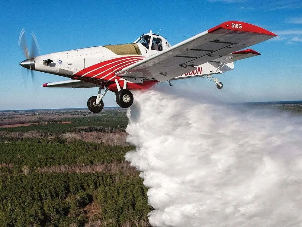 A white and red Thrush 510G crop duster aircraft in flight, spraying a white substance over a green forest.