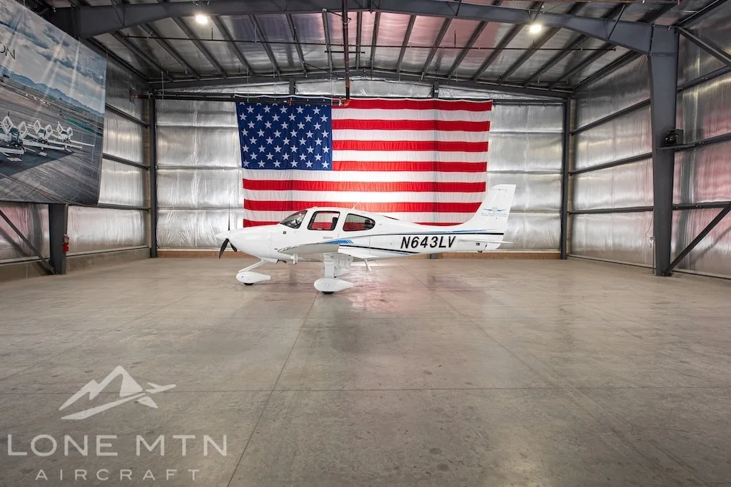 A white Cirrus SR22 aircraft with blue and black stripes, tail number N643LV, parked in a hangar with an American flag in the background.