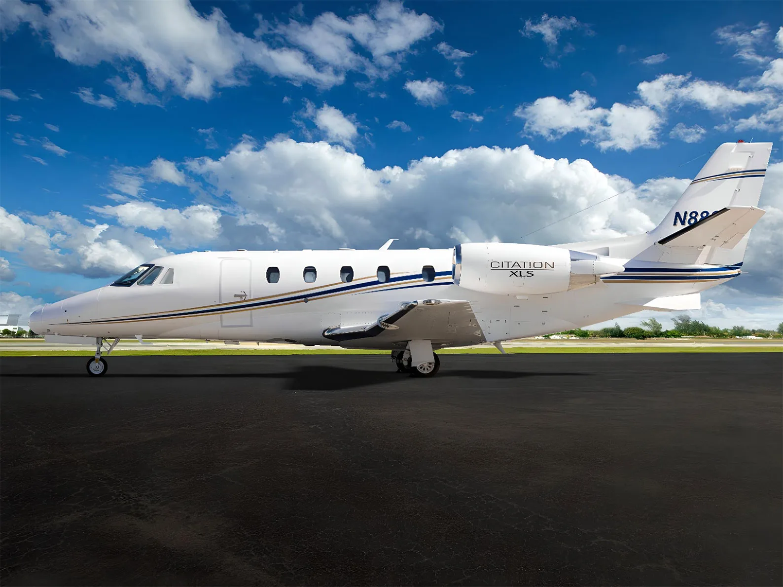 A white Cessna Citation XLS private jet with blue and gold stripes on the tarmac under a blue sky.