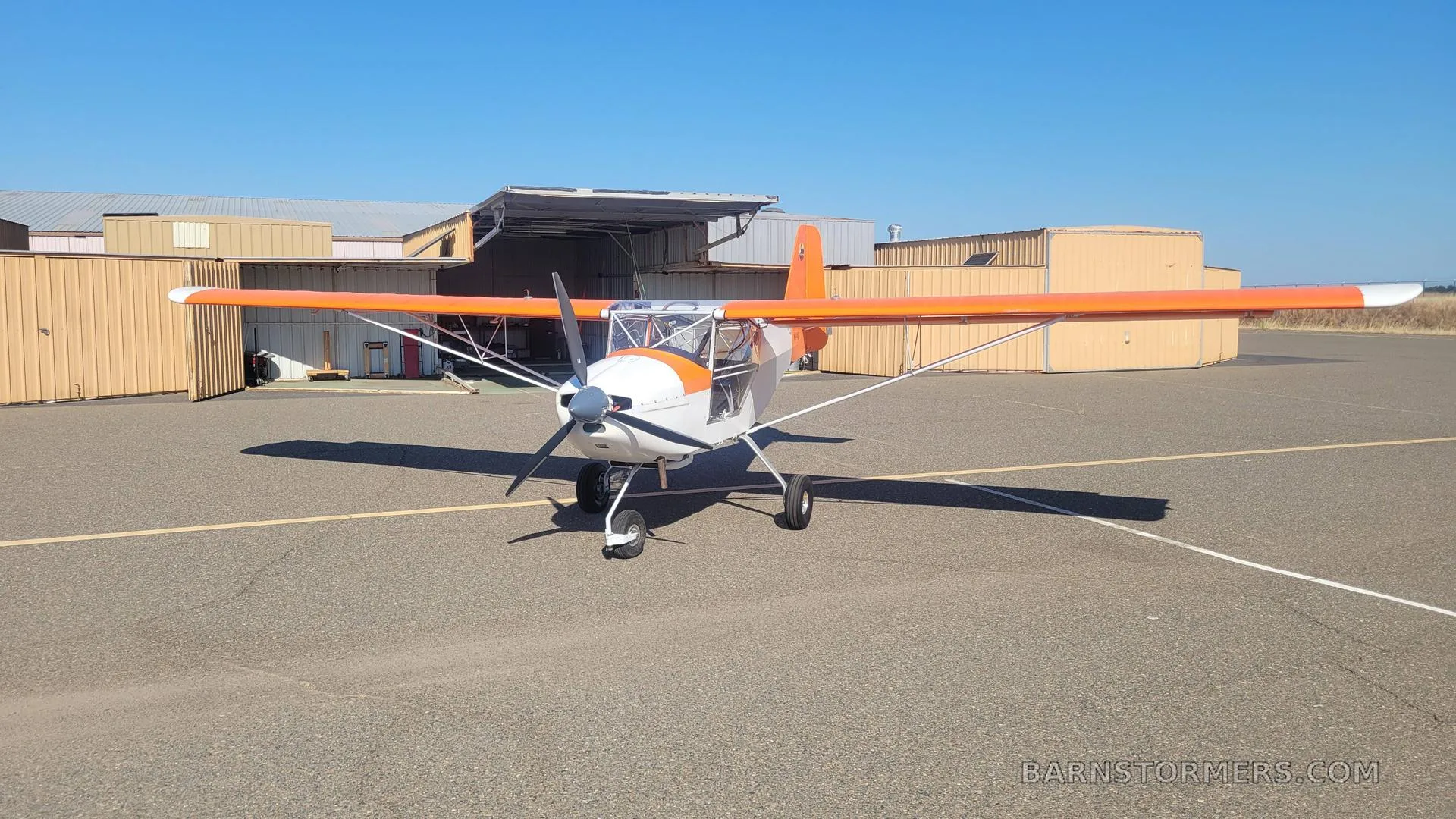 A white and orange high-wing aircraft with a propeller, parked on an asphalt apron in front of hangars under a clear blue sky.