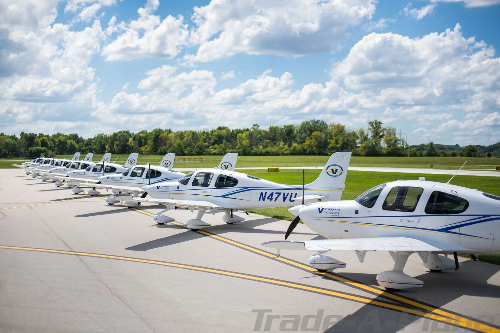 A line of white single-engine propeller aircraft, including N47VU, with blue and yellow stripes, parked on an airfield under a partly cloudy sky.